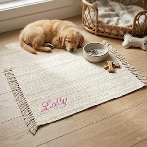 May include: A rectangular off-white woven mat featuring fringe on two ends and the name Lolly embroidered in pink script. The mat is placed on a wooden floor alongside a ceramic bowl containing dried lavender and a bone-shaped dog treat.