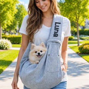 May include: A light gray pet carrier with the name "Luna" embroidered on the strap. A small, light-colored dog is nestled inside the carrier. The carrier is being worn by a person walking outside on a sunny day.