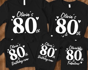 May include: Black t-shirts and a onesie with white text that reads "Olivia's 80th" and "Birthday crew". One shirt says "Olivia's 80th & Fabulous". The shirts have a heart design. The shirts are for a birthday.