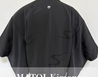 May include: Black kimono with a subtle pattern and a small emblem near the collar. The garment has wide sleeves and a draped design. The text "MATOI Kimono" is displayed at the bottom of the image.