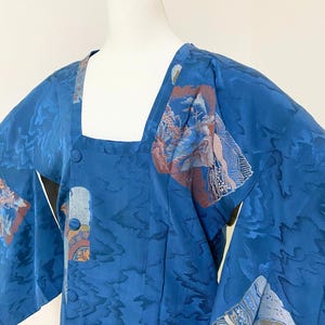 Könnte beinhalten: Blauer Kimono-Stil-Roben mit quadratischem Ausschnitt und drei Knöpfen. Die Robe hat ein strukturiertes Muster und Patches mit detaillierten Kunstwerken in Blau-, Braun- und Weißtönen. Die Ärmel sind weit und der Stoff scheint Seide zu sein.
