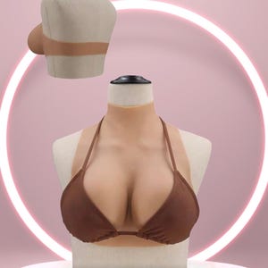 May include: A brown bikini top and a neck and chest prosthetic are displayed on a mannequin. The prosthetic is flesh-toned with a brown bikini top. The background is pink with a white neon circle.