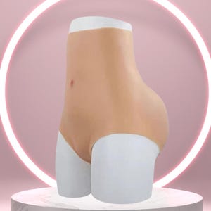 Pode incluir: Um modelador de corpo de silicone cor de pele com shorts brancos, exibido em um pedestal de mármore sobre um fundo rosa. O modelador de corpo tem uma aparência realista, com cintura e nádegas definidas.
