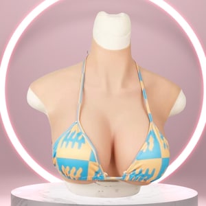 May include: A realistic silicone breast form with a halter-style bikini top. The bikini top features a blue and yellow checkered pattern. The form is displayed on a white pedestal against a pink background with a neon ring.