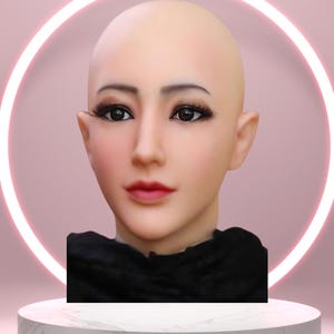 Op de afbeelding: Een realistisch siliconen masker van een vrouwelijk gezicht, met gedetailleerde gelaatstrekken zoals donkere ogen, lange wimpers en rode lippenstift. Het masker is kaal en heeft een gladde huidskleur. Het staat tegen een roze achtergrond met een neonring en een witte sokkel.