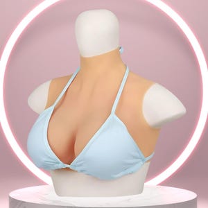May include: A light blue bikini top is displayed on a mannequin with a realistic skin-tone chest and shoulders. The mannequin is on a white pedestal against a pink background with a glowing circular light.