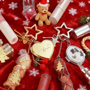 Puede incluir: Una disposición festiva de productos de belleza y accesorios sobre un fondo rojo. Incluye barras de labios, llaveros, un osito de peluche con gorro de Papá Noel y un relicario en forma de corazón. Decorado con copos de nieve.