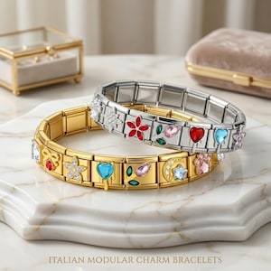 May include: Two Italian modular charm bracelets. One is gold-coloured, the other silver-coloured. Both feature colourful charms with floral, heart, and gemstone designs. The bracelets are displayed on a marble surface.