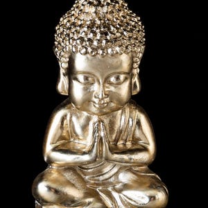 Könnte beinhalten: Eine metallisch goldfarbene Buddha-Figur in sitzender Haltung mit einer glatten, reflektierenden Oberfläche. Die Figur ist mit gekreuzten Beinen und zusammengelegten Händen in einer Gebetshaltung dargestellt, inklusive detaillierter Locken.