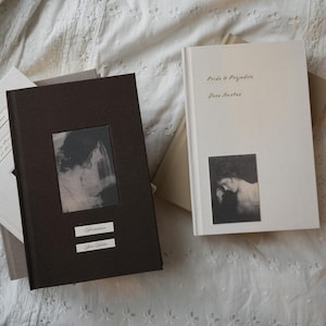Puede incluir: Dos libros de tapa dura, uno marrón y otro blanco, con imágenes y texto en blanco y negro. El libro marrón tiene el título "Persuasión" y el nombre del autor. El libro blanco se titula "Orgullo y Prejuicio" de Jane Austen.
