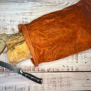 May include: A loaf of bread and a slice of bread are next to an orange bread bag with a leaf pattern. A serrated bread knife with a black handle is on a white wooden surface.