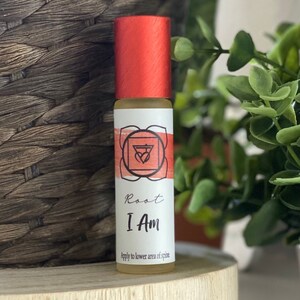 Puede incluir: Un frasco de roll-on rojo y blanco con una tapa roja. La etiqueta dice "Root I Am" y "Aplicar en la zona inferior de la columna vertebral".