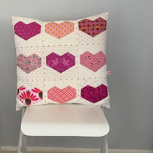 May include: A white pillow with a patchwork design of pink and purple hearts. The pillow is sitting on a white chair.