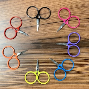 May include: A set of eight small scissors with colourful handles. The handles are black, red, pink, purple, blue, and yellow. The scissors are arranged in a circle on a wooden surface.
