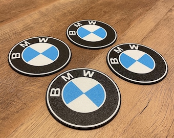 Puede incluir: Cuatro posavasos redondos con el logotipo de BMW. Cada posavasos tiene un borde negro, un centro blanco con una cruz azul y las letras "BMW". Los posavasos están sobre una superficie de madera.
