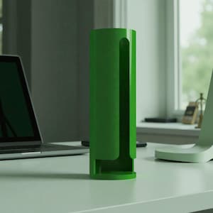 Op de afbeelding: Een hoge, cilindervormige, heldergroene bureau-organizer met een gebogen sleuf aan één kant. De organizer is gemaakt van een stevig materiaal en staat op een witte tafel. Een laptop en een computer zijn op de achtergrond te zien.