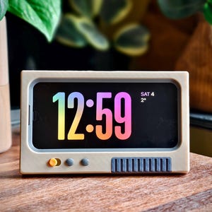 Puede incluir: Un reloj blanco de estilo retro con una pantalla negra que muestra la hora 12:59 y la fecha SÁB 4 2°. El reloj tiene tres botones redondos y una fila de botones rectangulares.
