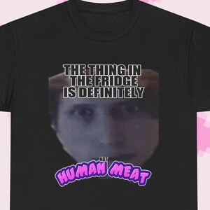 May include: Black t-shirt with a graphic featuring a person's face and the text "THE THING IN THE FRIDGE IS DEFINITELY NOT HUMAN MEAT" in white and purple. The shirt is a basic crew neck style.
