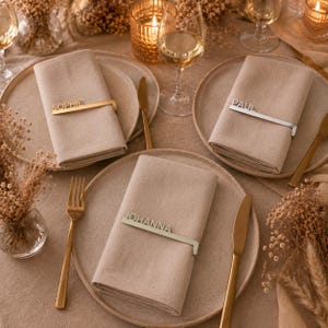 Può includere: Un'elegante mise en place con tovaglioli di lino beige, ciascuno tenuto da un portanome in metallo. I nomi "SOPHIE", "PAUL" e "JOHANNA" sono visibili. Posate dorate e argentate, piatti, candele e bicchieri di vino completano l'allestimento.