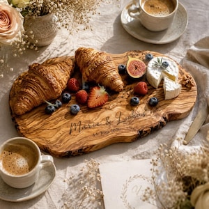 Pode incluir: Uma t&aacute;bua de servir de madeira com croissants, frutos silvestres, queijo e um figo. A t&aacute;bua est&aacute; gravada com os nomes "Marie & Lukas" e a data "14. Juni 2026". Tamb&eacute;m est&atilde;o presentes duas ch&aacute;venas de caf&eacute;.
