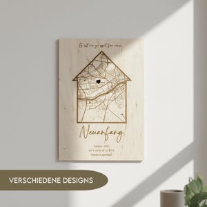 Op de afbeelding: Houten wandkunst met een huisvormige kaart van een stad, gemarkeerd met een hart. Het woord "Neuanfang" staat onder de kaart, samen met coördinaten en de naam van de stad. De tekst "VERSCHIEDENE DESIGNS" staat onderaan.