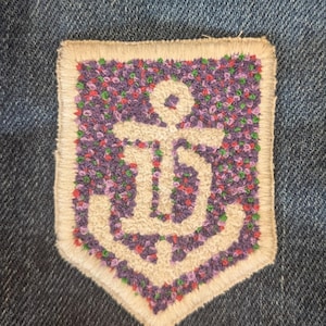 May include: Embroidered patch featuring a white anchor design on a purple floral background. The patch has a cream-colored border and is set against a blue denim fabric. The anchor is a nautical symbol.