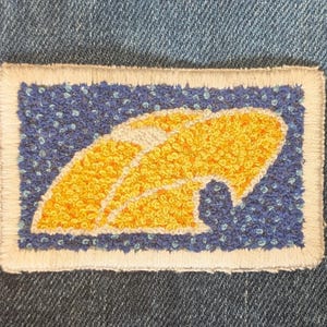 Op de afbeelding: Geborduurde patch met een geel en wit ontwerp op een blauwe achtergrond. De patch heeft een witte rand en is rechthoekig. De patch staat op een denim achtergrond.
