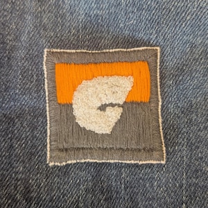May include: Close-up of a denim fabric with a square patch. The patch features a gray border, an orange center, and a white, textured letter "G". The patch is sewn onto the blue denim.