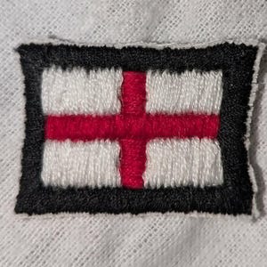 Op de afbeelding: Een rechthoekige geborduurde patch met de vlag van Engeland, bestaande uit een rood kruis op een witte achtergrond. Het gehele ontwerp is omrand door een dikke, effen zwarte geborduurde rand, wat zorgt voor een duidelijk kader.