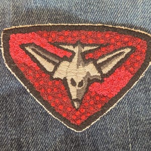 May include: Embroidered patch with a shield shape, featuring a gray stylized aircraft design on a red background. The patch is outlined in black and white, set against a blue denim fabric.
