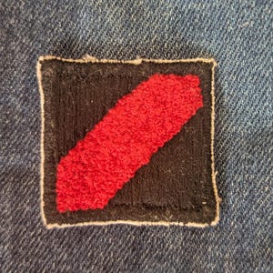 May include: A square patch with a black background and a diagonal red stripe. The patch is sewn onto a blue denim fabric. The red stripe has a textured, raised appearance, and the patch is outlined with a white border.