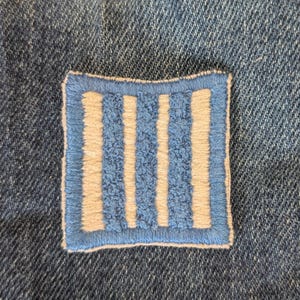 May include: A square patch with a blue border and vertical stripes. The stripes alternate between light beige and blue, creating a textured appearance. The patch is sewn onto a denim fabric, providing a contrast in textures and colors.