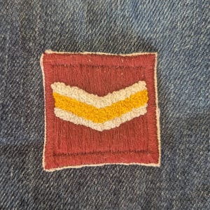 May include: A maroon and yellow chevron patch, stitched onto a blue denim fabric. The patch has a square shape with a maroon border and a yellow and white chevron design in the center. The denim fabric has a textured appearance.