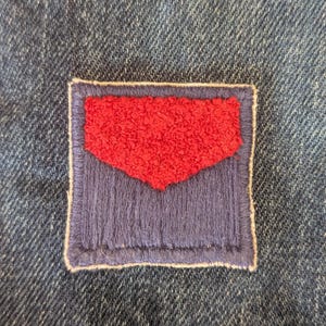 May include: Close-up of denim fabric with a stitched patch. The patch has a red textured design within a purple border, outlined with white stitching. The denim has a blue wash.