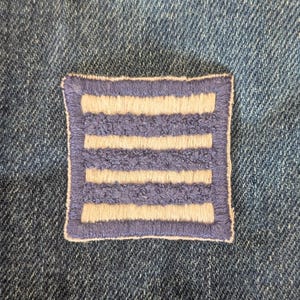 May include: A square patch with a light blue border and four horizontal cream-colored stripes. The patch is set against a denim background, creating a contrast in textures and colors. The patch is likely an embroidered emblem.