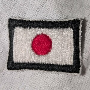 May include: A rectangular embroidered patch featuring a white background with a solid red circle in the centre, enclosed by a thick black stitched border. The embroidery shows visible thread texture throughout the design on a white fabric base.