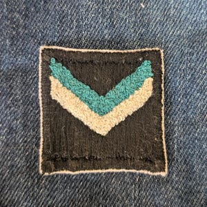 May include: A square, black patch with a teal and off-white chevron design. The patch is sewn onto a blue denim fabric, with the chevron pattern centered within the black square.