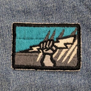 May include: Embroidered patch featuring a hand grasping a lightning bolt design. The patch has a teal background with gray and white accents, and a black border. The patch is rectangular and appears to be sewn onto a denim fabric.