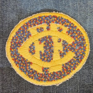 May include: Embroidered patch with a yellow circular design and the letters "JFC" in the center. The patch has a blue, red, and yellow speckled border and is set against a denim background.