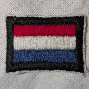 May include: A rectangular embroidered patch featuring a horizontal tricolor design of red, white, and blue stripes. The entire flag motif is enclosed within a thick, solid black stitched border, creating a distinct frame around the colored threads.