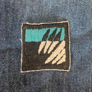 Peut inclure: Un écusson carré brodé avec une bordure et un fond noirs. L'écusson présente une bande horizontale turquoise en haut et trois formes blanches en forme de griffes sur la moitié inférieure. L'écusson est cousu sur un tissu en denim bleu.
