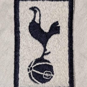 May include: A rectangular embroidered patch featuring a dark navy blue cockerel standing atop a dark navy blue football on a light cream background. The patch is bordered by a thick, dark navy blue stitched frame, creating a clean, defined edge.