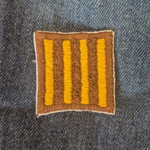 May include: A square patch with a brown border and a brown interior featuring four vertical yellow stripes. The patch is sewn onto a blue denim fabric, creating a contrast in textures and colors.