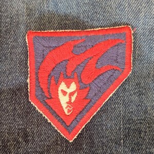 May include: Embroidered patch with a red devil design on a purple background, bordered in red. The patch features a white face and horns, and is sewn onto denim fabric.