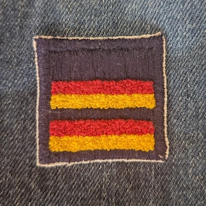 May include: A rectangular patch with a navy blue background and two horizontal stripes in red and yellow. The patch is sewn onto a denim fabric, creating a contrast in textures and colors. The patch has a white border.