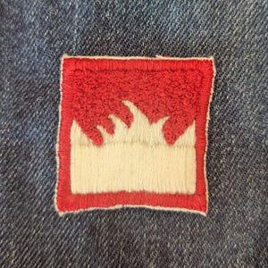 May include: A square, embroidered patch with a red border and a white and red flame design. The patch is sewn onto a blue denim fabric, creating a contrast in textures and colors. The design is simple yet striking.