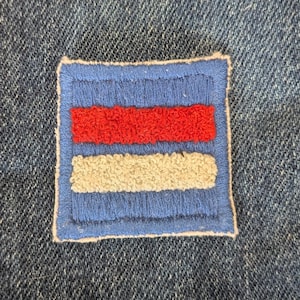 May include: A square patch with a blue border and background, featuring a red horizontal stripe above a white horizontal stripe. The patch is sewn onto a denim fabric.