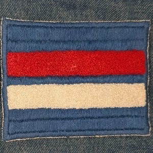 May include: A rectangular embroidered patch featuring a blue background with a horizontal red stripe and a horizontal white stripe. The patch is stitched onto a blue denim fabric base, showcasing a textured, raised thread design throughout.
