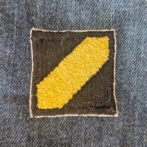 May include: A square patch with a black background and a diagonal yellow stripe. The patch is sewn onto a blue denim fabric. The yellow stripe has a textured, raised appearance, and the patch is outlined with a white border.