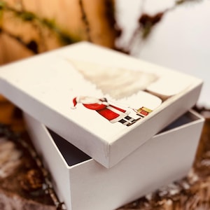 Könnte beinhalten: Eine rechteckige weiße Geschenkbox mit einem Deckel, der eine gedruckte Illustration des Weihnachtsmanns im roten Anzug neben einem stilisierten Weihnachtsbaum zeigt. Die Box ist geöffnet und zeigt das weiße Innere sowie den schräg aufliegenden Deckel.
